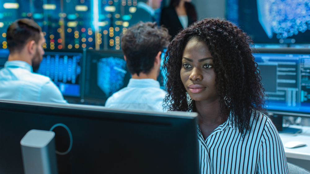 IT professional in data center looks at her monitor, searching for cyber security issues, as two colleagues work in background. 