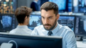 IT professional in data center looks at monitor, searching for Log4j issues, as colleague works at row of monitors behind him. 